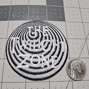 May include: A circular embroidered patch featuring a black and white spiral pattern with the text THE TWILIGHT ZONE stitched in white across the center. A United States quarter coin is placed next to the patch for a clear size reference.