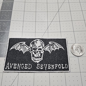 May include: A rectangular black embroidered patch featuring a white skull with bat wings and the text "AVENGED SEVENFOLD" in a stylized font. A US quarter coin is placed to the right of the patch for scale, showing the relative size of the item.
