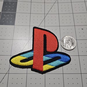 May include: An embroidered patch featuring the PlayStation logo with a red vertical bar and a base composed of yellow, light blue, and dark blue segments, outlined in black, positioned next to a United States quarter for scale reference.