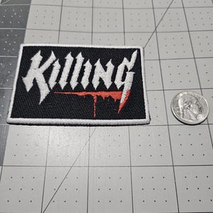May include: A rectangular embroidered patch with a black background and a white border. The word "Killing" is stitched in a stylized white font across the center, with a red dripping effect underneath the letters, positioned next to a US quarter.