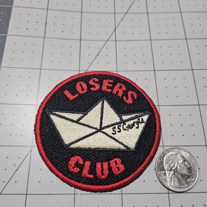 May include: A circular embroidered patch with a black background and a red border. The patch features a white paper boat with the text SS Georgie on its side, and the red text LOSERS CLUB arched above and below the boat, next to a US quarter.