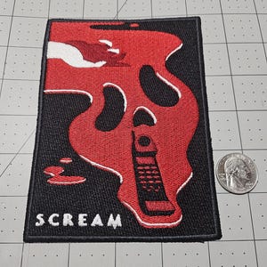 May include: A rectangular embroidered patch featuring a black background with a red, melting ghost-like face design. The design includes a white hand, a black vintage mobile phone, and the word SCREAM in white text. A US quarter is shown for scale.