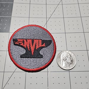 May include: A circular embroidered patch featuring a black anvil silhouette with the red text ANVIL across the center. The patch has a grey background and a red border. A US quarter coin is placed next to the patch for scale reference.
