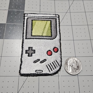 May include: An embroidered patch shaped like a handheld video game console in light gray with a lime green screen, a black directional pad, two red circular buttons, and black speaker slits, positioned next to a US quarter for scale reference.