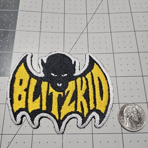 May include: A black and yellow embroidered patch shaped like a bat with the text BLITZKID in yellow letters. The patch is positioned next to a US quarter coin for scale, showing the patch is approximately 3.5 inches wide by 2.5 inches tall.
