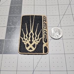 May include: A rectangular embroidered patch with a black background and gold stitching. The design features a stylized graphic symbol and the text "Static-X" in a vertical orientation. A US quarter coin is placed next to the patch for scale.
