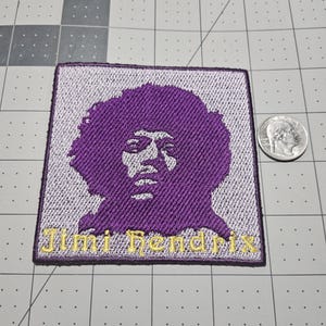 May include: A square embroidered patch featuring a purple silhouette portrait of Jimi Hendrix on a light purple background. The name Jimi Hendrix is stitched in gold thread at the bottom. A US quarter coin is placed next to the patch for scale.