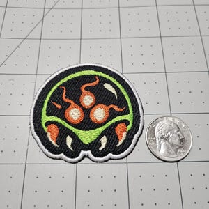May include: A circular embroidered patch featuring a black background with a lime green border. The design includes three orange circular shapes with tails and two orange claw-like elements at the bottom. A US quarter is placed next to the patch for scale.