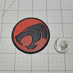 May include: A circular embroidered patch featuring a black stylized cat head logo on a red background, with a black border. The patch is positioned next to a United States quarter coin for scale, providing a clear visual reference for its size.