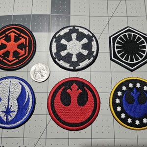 May include: Six embroidered fabric patches featuring various circular and hexagonal designs in red, black, white, blue, and yellow. A silver United States quarter coin is placed between the patches for scale, measuring approximately 0.955 inches in diameter.