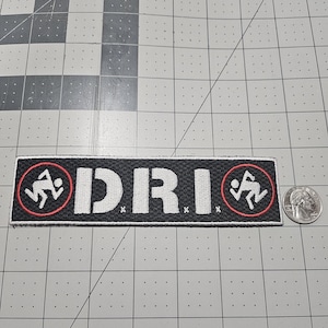 May include: A rectangular embroidered patch featuring the white text D.R.I. flanked by two circular logos with white running figures inside red rings on a black background. A US quarter coin is placed next to the patch for scale reference.