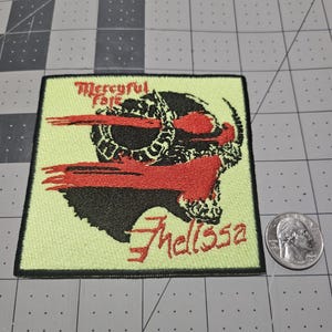 May include: A square embroidered patch featuring a pale yellow background with a black and red graphic of a horned skull. The text Mercyful Fate and Melissa is stitched in red. A US quarter is placed next to the patch for scale reference.