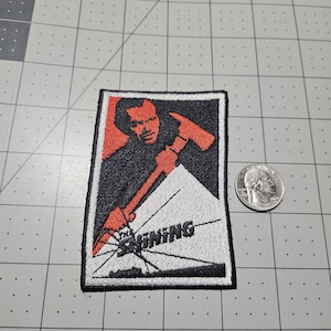May include: A rectangular embroidered patch featuring a graphic of a person holding an axe, with the text THE SHINING printed over a cracked white background. A US quarter coin is placed next to the patch for scale on a grid mat.