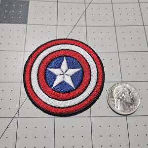 May include: A circular embroidered patch featuring a central white star on a blue background, surrounded by concentric rings of white and red, with a black border. A United States quarter coin is placed next to the patch for size reference.