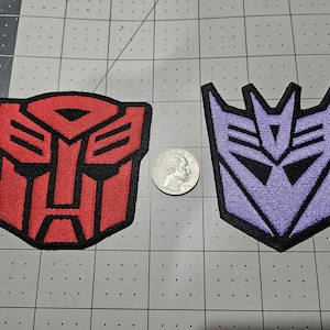 May include: Two embroidered patches featuring the Autobot and Decepticon logos. The red Autobot patch and the purple Decepticon patch are positioned on either side of a silver United States quarter coin for scale, showing their relative size.