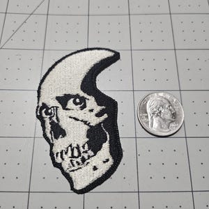 May include: A black and white embroidered patch featuring a stylized skull design with a crescent moon shape. The patch is positioned next to a United States quarter coin for scale, showing the relative size of the embroidered textile item.