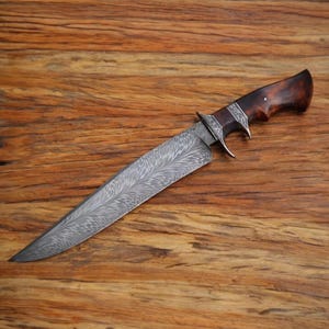 Puede incluir: Un cuchillo artesanal con una hoja de acero estampada y un mango de madera oscura. La hoja tiene un diseño intrincado y grabado. El mango es de un marrón oscuro intenso con una forma curvada. El cuchillo se muestra sobre una superficie de madera.