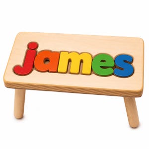 Puede incluir: Un taburete pequeño de madera con un acabado natural claro que presenta un recorte en la superficie superior. El recorte contiene cinco letras de madera individuales que deletrean el nombre "james" en colores rojo, naranja, amarillo, verde y azul.