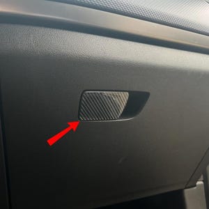 Op de afbeelding: Een close-up van een zwart auto dashboard met een carbon fiber textuur handschoenenkastje handvat. De handgreep is rechthoekig met afgeronde hoeken en een textuurpatroon. Het dashboard heeft een textuuroppervlak.