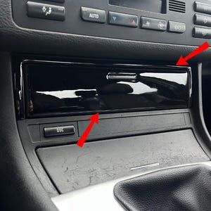 Op de afbeelding: Een glanzend zwart opbergvak voor de console van de auto. Het vak is rechthoekig met een kleine handgreep en bevindt zich onder de bedieningselementen van het dashboard. De DSC-knop is eronder zichtbaar.