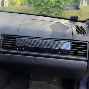 May include: A black car dashboard featuring a central carbon fiber trim insert positioned between two air vents. The dashboard surface has a textured finish, and the passenger side includes an SRS airbag emblem embossed on the upper panel.