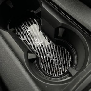 Può includere: Un inserto per portabicchieri per auto con motivo in fibra di carbonio e la scritta "NEXT OR LOGO" in colore chiaro. L'inserto è progettato per adattarsi perfettamente all'interno del portabicchieri di un'auto, offrendo un aggiornamento elegante e funzionale.