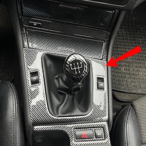 Puede incluir: Consola interior de coche con un patrón de fibra de carbono. La imagen muestra una palanca de cambios, controles de ventanas y otros botones. La consola es negra y gris, con asientos de cuero visibles.
