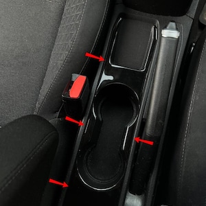 Pode incluir: Console de carro preta com porta-copos, compartimento de armazenamento e travão de mão. A consola é rodeada por bancos de carro de tecido preto e fivelas de cinto de segurança vermelhas. A consola tem um acabamento brilhante.