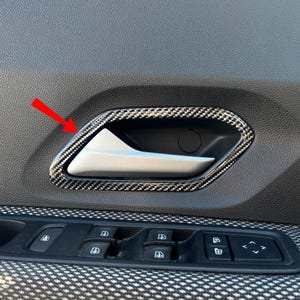 Può includere: Una maniglia interna per portiera auto con leva argentata inserita in una cornice con motivo in fibra di carbonio. Una freccia rossa indica l'angolo superiore sinistro della finitura. La maniglia è posizionata sopra un pannello finestrini.