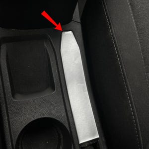 Può includere: Un ricevitore per fibbia della cintura di sicurezza per auto, di colore argento e di forma rettangolare, è posizionato tra la console centrale e il sedile del passeggero. L'interno circostante è nero.