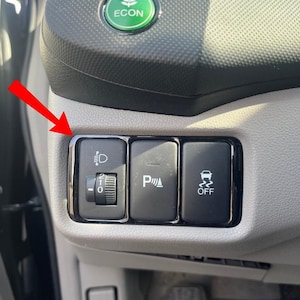 Può includere: Primo piano del cruscotto di un'auto con vari pulsanti di controllo. Sono inclusi i comandi dei fari, l'assistenza al parcheggio e un interruttore di disattivazione del controllo di stabilità. Il cruscotto è grigio e nero.