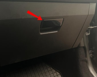 Op de afbeelding: Een zwart dashboardkastje met een rechthoekige handgreep. Het compartiment is gesloten en lijkt van plastic te zijn. De binnenkant van de auto is donker, met een rubberen vloermat zichtbaar.