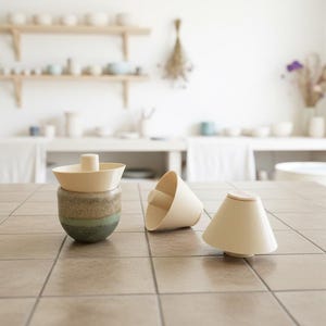 Può includere: Un insieme di pezzi in ceramica fatti a mano, tra cui uno spremiagrumi color crema e due piccoli vasi a forma di cono. Lo spremiagrumi è posto su una base smaltata multicolore. Gli oggetti sono disposti su una superficie piastrellata.