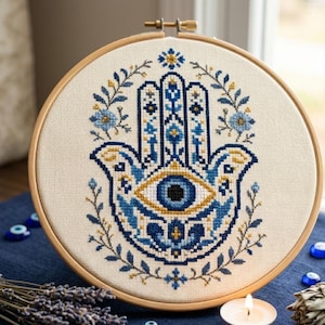 May include: A cross-stitch embroidery of a Hamsa hand with an eye in the center, framed in a wooden hoop. The design features blue and gold detailing on a cream-colored background, surrounded by floral accents. The embroidery is approximately 8 inches in diameter.