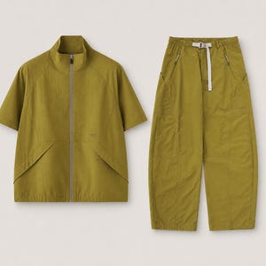 Könnte beinhalten: Ein zweiteiliges Outfit in Olivgrün. Das Oberteil ist eine kurzärmelige Jacke mit hohem Kragen und durchgehendem Reißverschluss. Die Hose ist weit geschnitten mit einem Kordelzug in der Taille. Das Outfit ist aus leichtem Stoff, möglicherweise Nylon.