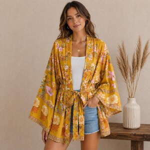 Könnte beinhalten: Senfgelber Kimono mit floralem Print und Bindegürtel. Der Kimono hat ein lebendiges Blumenmuster in Rosa-, Orange- und Blautönen. Die Ärmel sind weit und fließend, und das Kleidungsstück ist aus leichtem Stoff gefertigt.
