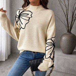 Könnte beinhalten: Cremefarbener Pullover mit Stehkragen und langen Ärmeln. Der Pullover hat ein schwarzes Blumendesign auf der Brust und am Ärmel. Die Person trägt eine blaue Jeans und hält eine schwarze Handtasche.