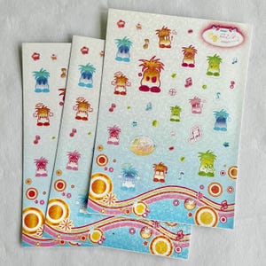 May include: Sheets of stickers with cartoon characters in blue, orange, and green. The stickers are on a light blue background with musical notes and decorative circles. The stickers have various hairstyles.