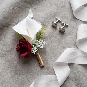 May include: A boutonniere featuring a white calla lily, a deep red rose, and small white filler flowers with green leaves, wrapped in gold ribbon, accompanied by a pair of silver-toned metal cufflinks and a long white ribbon.