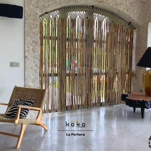 May include: Interior scene featuring a woven chair with a zebra-striped pillow, a wooden table with a gold lamp, and a macrame curtain. The room has a neutral color palette with natural textures and the text "koko La Portera".