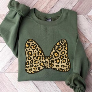 May include: An olive green sweatshirt featuring a large bow design with a leopard print pattern in shades of tan and black. The sweatshirt has long sleeves and a crew neck. The bow is centered on the front of the garment.