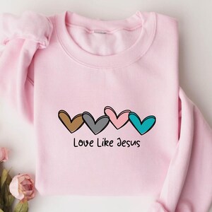 May include: A light pink sweatshirt featuring a graphic of four hearts in brown, gray, pink, and teal. Below the hearts is the text "Love Like Jesus" in black script. The sweatshirt has long sleeves and a crew neck.