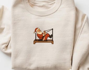 May include: Cream-colored sweatshirt featuring a cartoon Santa Claus exercising on a Pilates reformer machine. Santa is depicted in his traditional red suit and hat. The design is centered on the chest of the sweatshirt.
