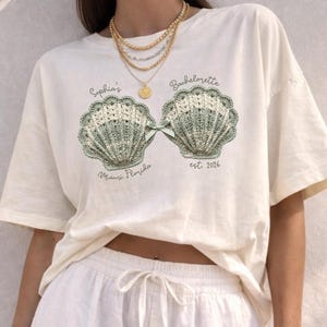 May include: Off-white t-shirt featuring a seashell design with the text "Sophia's Bachelorette" and "est. 2026". The design includes a green bow and the words "Miami Florida". The shirt is a casual style.
