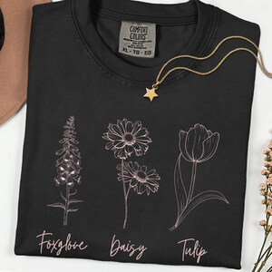 May include: Black Comfort Colors t-shirt with a floral design featuring a foxglove, daisy, and tulip. The names of the flowers are written in a cursive font below each flower. A gold star pendant necklace is draped on the shirt.