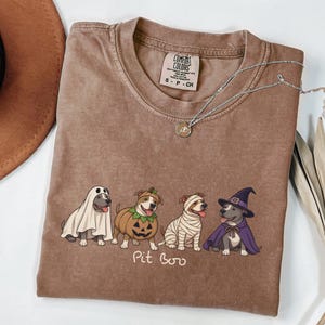 May include: A brown short-sleeve t-shirt featuring a graphic of four dogs in Halloween costumes: a ghost, a pumpkin, a mummy, and a witch. Below the dogs, the text reads Pit Boo. The shirt has a crew neckline and a Comfort Colors brand label.