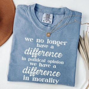 May include: A blue Comfort Colors t-shirt with the text "we no longer have a difference in political opinion we have a difference in morality" in white. A gold necklace with a small pendant is on the shirt.