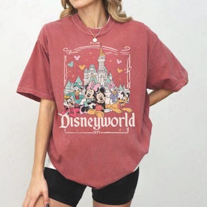 May include: A maroon-colored t-shirt featuring a Disneyworld graphic. The graphic includes a castle, Mickey and Minnie Mouse, Donald Duck, Daisy Duck, and Goofy. The text "Disneyworld" is displayed prominently, with "1971" below.
