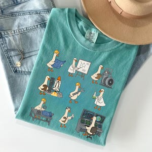 May include: Teal Comfort Colors t-shirt featuring cartoon geese in various engineering and scientific roles, including a blueprint, a whiteboard with a chemical formula, and a rocket launch. The shirt is paired with light wash jeans and a tan hat.