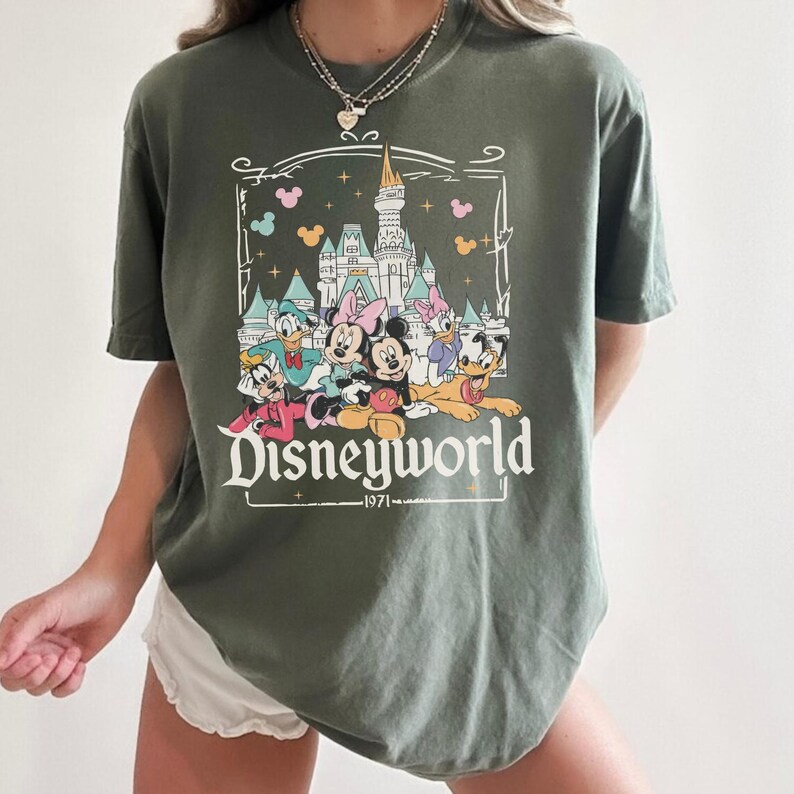 May include: Olive green t-shirt featuring a Disneyworld graphic. The design includes a castle, Mickey and Minnie Mouse, Donald Duck, Daisy Duck, Goofy, and Pluto. The text "Disneyworld" and "1971" are also included in the design.